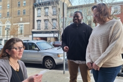 Lynn Alexander sits and reads a poem aloud outside as a man and woman stand and listen.