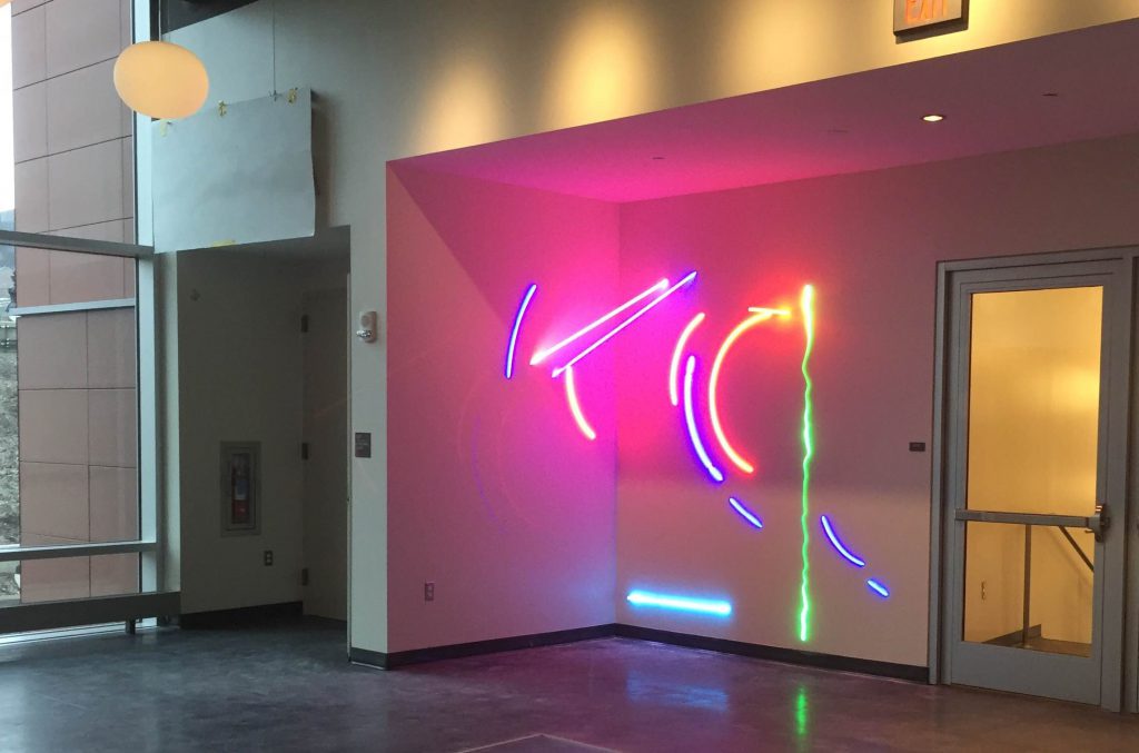 The sculpture For JT by Stephen Antonakas on the Karl Stirner Arts Trail features blue, orange, and light green neon tubing- on a wall in Buck Hall, Lafayette College.