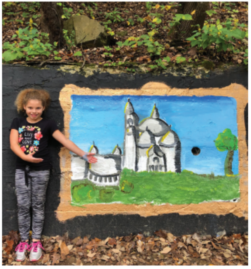 A young girl stands by and points to her painting of a building, possibly the Taj Mahal, on the Young Masters Wall on the Karl Stirner Arts Trail in Easton, Pennsylvania.