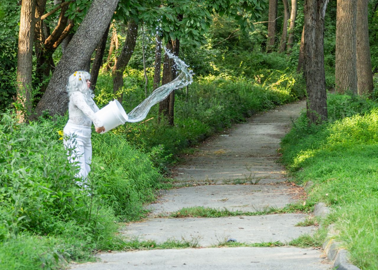 A woman in white tosses water from a bucket onto the Karl Stirner Arts Trail in a performance event held in conjunction with the Upriver artwork by Heidi Wiren Bartlett.