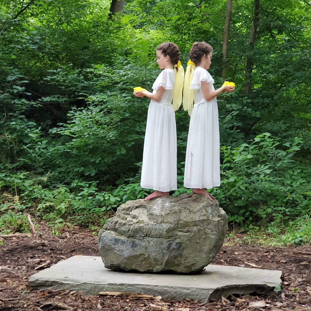 Two girls dressed in white holding yellow flowers stand on top of a Stromatolite rock during an event for the Upriver exhibition by Heidi Miren Bartlett at the Karl Stirner Arts Trail in Easton, Pennsylvania. 