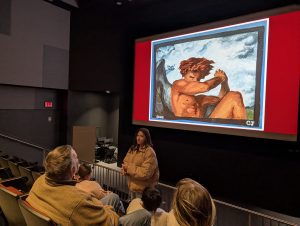 A young woman stands and talks to the audience about her Young Masters Wall painting of an angelic creature with wings as it's projected behind her during an event held by the Karl Stirner Arts Trail in Easton, Pennsylvania, in Lafayette College's Buck Hall.