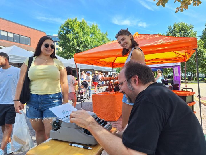 Two women smile as they watch poet Darrell Parry compose a spontaneous poem for them on an old-fashioned typewriter at the Easton Farmers' Market as part of the Karl Stirner Arts Trail's Poetry in the Wild Project.