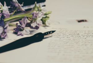 Poetry written in cursive on paper with a pen and purple flowers also on the paper