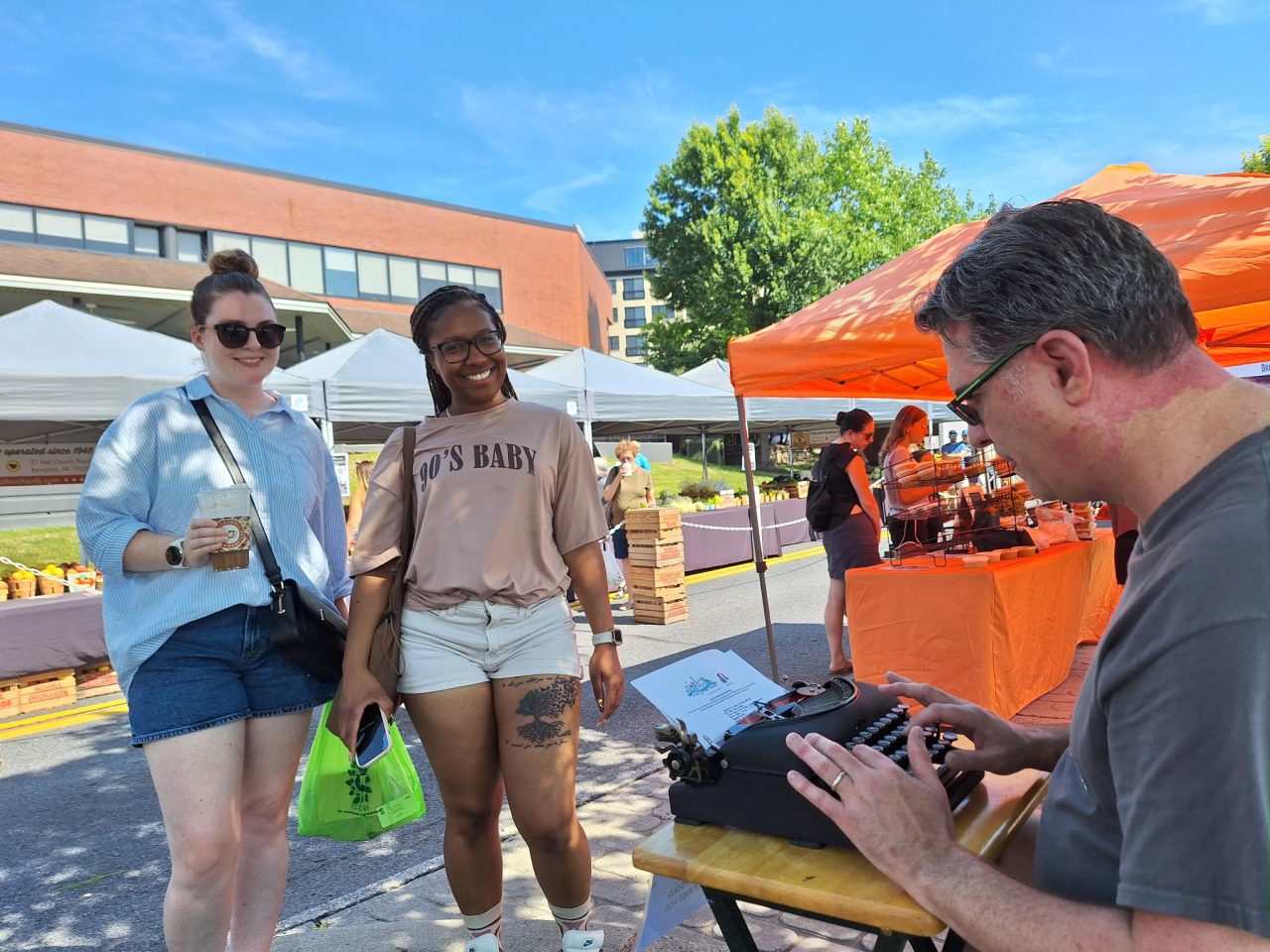 Two women smile while watching Matt Wolf type a spontaneous poem at the Easton Farmers Market in Easton, Pennsylvania, as part of the Karl Stirner Arts Trail's Poetry in the Wild project.