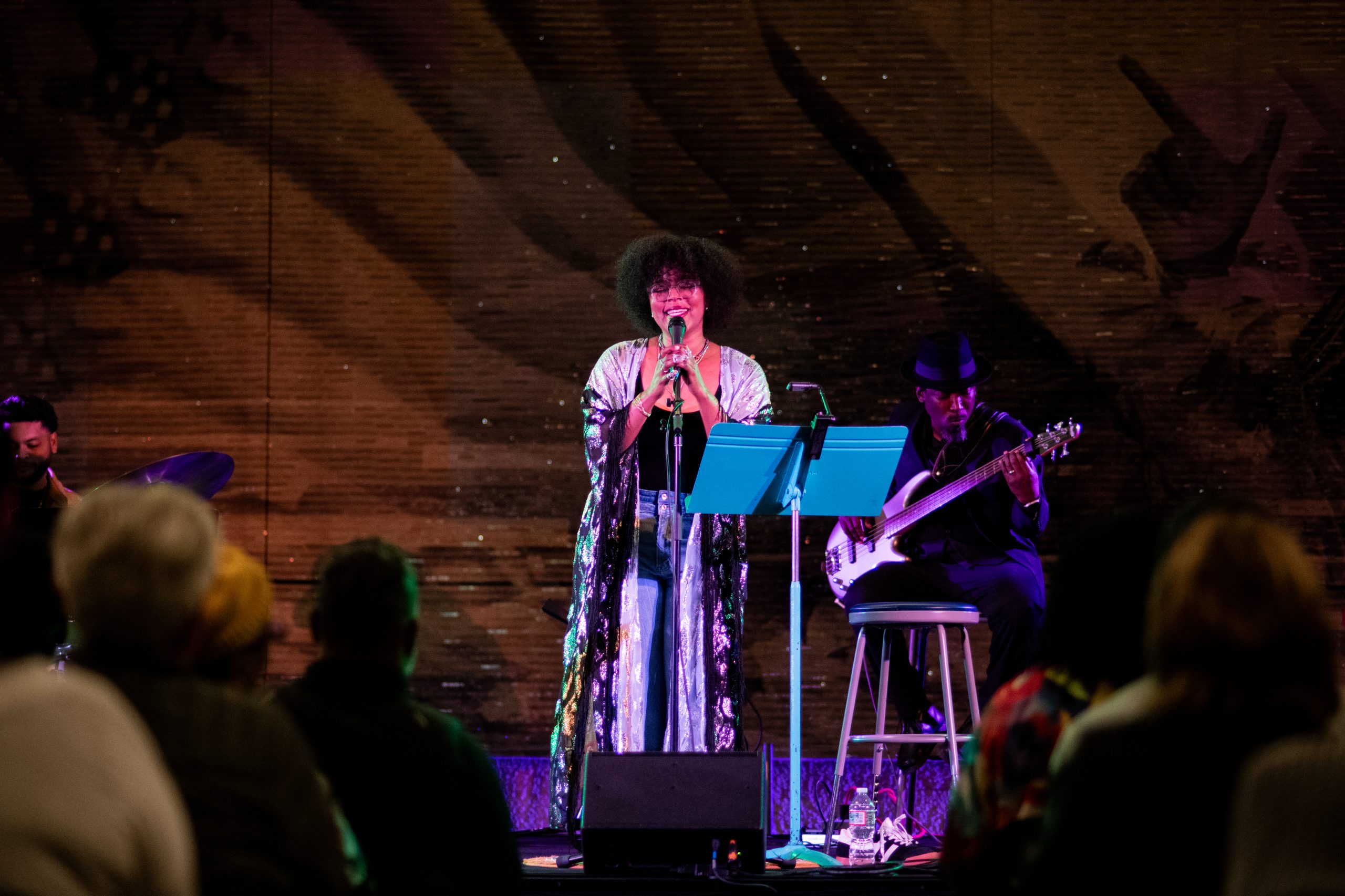 Yolanda Wisher sings at a microphone while a musician plays electric guitar before an audience at an Afro Eaters concert.