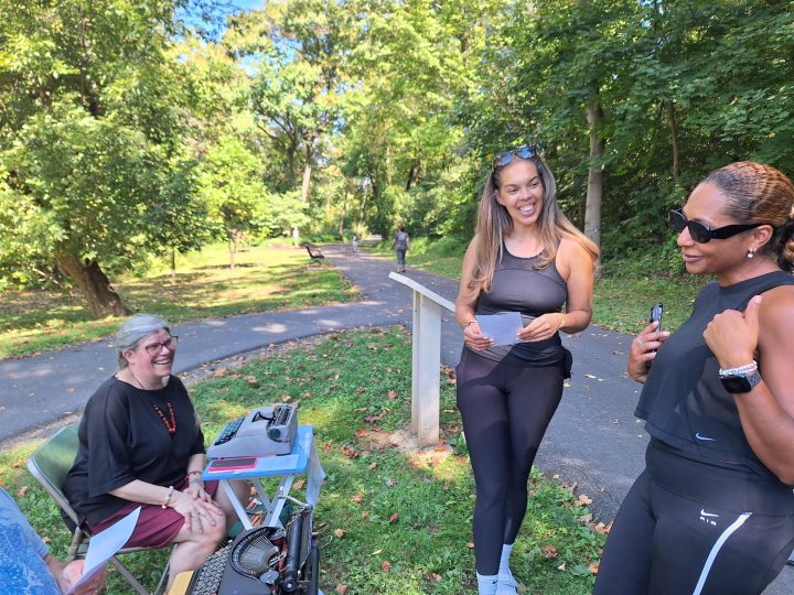 A woman holds a typed poem while talking with a woman friend as poet Rebecca Migdal sits at a typewriter on the Karl Stirner Arts Trail as part of its Poetry in the Wild Project.