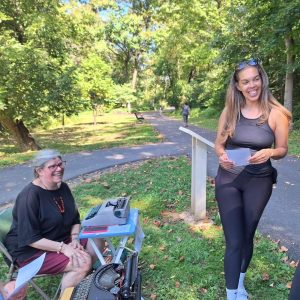 A woman holds a typed poem as poet Rebecca Migdal sits at a typewriter on the Karl Stirner Arts Trail as part of its Poetry in the Wild Project.